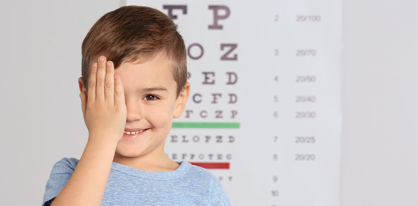 Ein Junge, der vor einer Wand mit einem Sehtest steht und sich ein Auge zuhält.