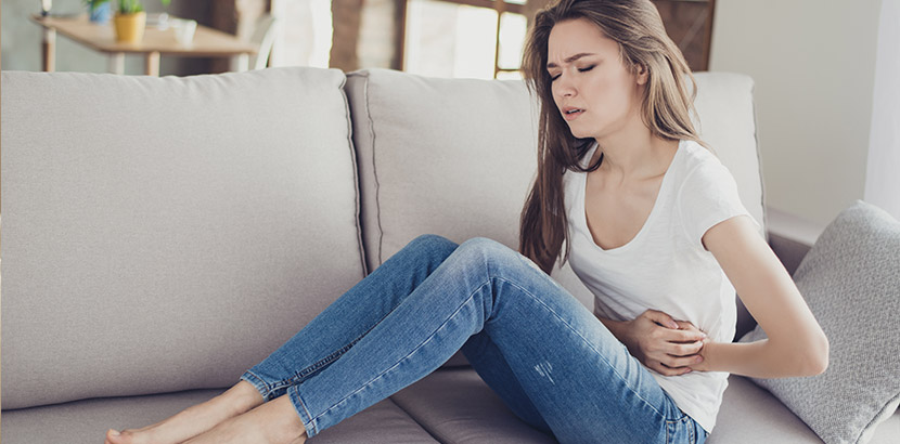 Junge hübsche Frau, die auf dem Sofa sitzt und sich wegen Unterleibsschmerzen krümmt. Endometriose Symptome.