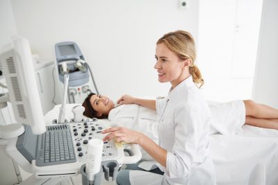 Blonde Endokrinologin, die die Schilddrüse einer brünetten Patientin ultraschallt. Schilddrüsenspezialist Wien.