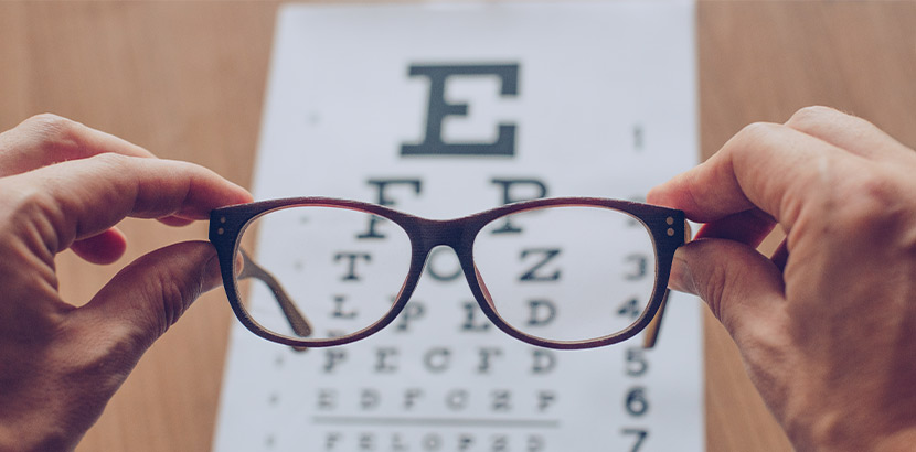 Zwei Hände, die eine braune Brille vor eine Sehtesttafel halten.