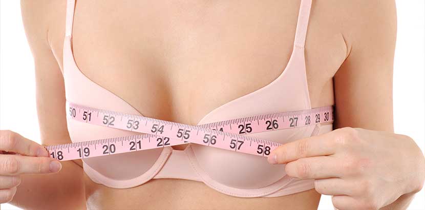 Oberkörper einer sehr schlanken Frau mit kleinen Brüsten in einem rosa BH, die ihre Brustgröße mit einem Maßband misst. Brustvergrößerung Wien Kosten.