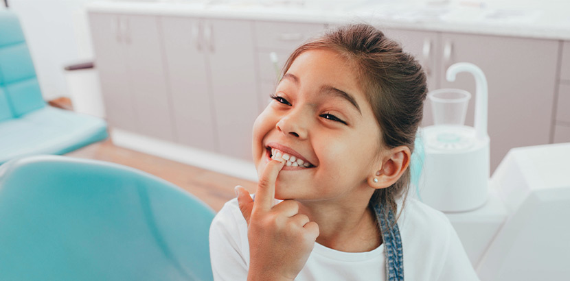 Ein Mädchen auf einem Zahnarztstuhl, das mit dem Finger auf ihre Zähne zeigt.