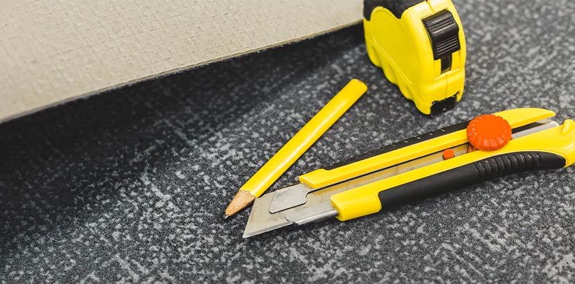 Ein Maßband, ein Bleistift und ein Stanleymesser liegen auf einem Stück Teppich.