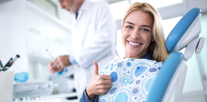Eine Frau im Zahnarztstuhl lächelt und zeigt Daumen hoch.
