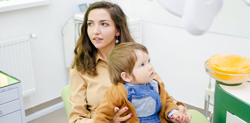 Eine Mutter sitzt mit ihrem Kind auf dem Schoß im Behandlungsraum eines Zahnarztes.