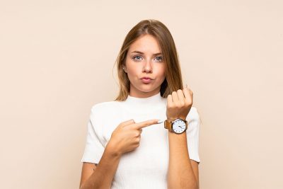 Junge Frau vor rosa Hintergrund, die mit einem ernsten Gesichtsausdruck auf ihre Armbanduhr zeigt. Pille danach Österreich.