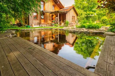 Ein Naturpool voller Wasser mit Haus und Terrasse aus Holz im Hintergrund.