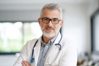 Ein Arzt mit grauem Haar und Brille lächelt.