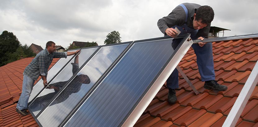 Zwei Installateure montieren Sonnenkollektoren für eine Solarthermieanlage mit Speicher auf einem Dach.