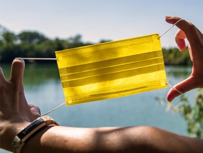 gelbe Maske, die eine Person von sich weghält, im Hintergrund ein See
