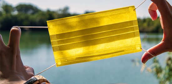 gelbe Maske, die eine Person von sich weghält, im Hintergrund ein See