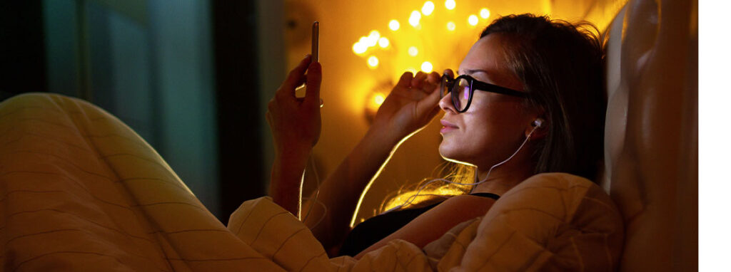 Junge Frau mit Brille, die nachts im Bett sitzt und via Smartphone etwas auf Google recherchiert. Corona Marketing oder was passiert nach Corona?