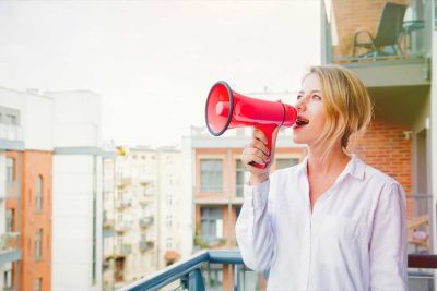 Junge blonde Frau, die mit einem Megafon auf ihrem Blakon steht und Werbung für ihre Website macht,