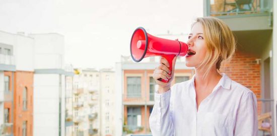Junge blonde Frau, die mit einem Megafon auf ihrem Blakon steht und Werbung für ihre Website macht,