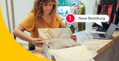 Modeverkäuferin, die ein Paket für den Versand einpackt. Neben ihr ein Laptop mit der Benachrichtigung, dass eine neue Bestellung eingegangen ist
